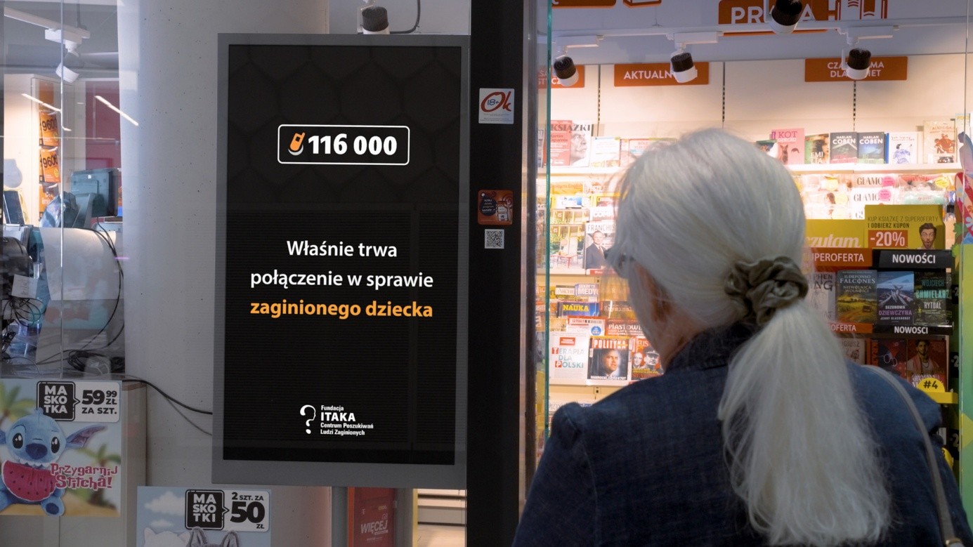 Widoczne wnętrze sklepu, a przed nim kobieta, kótra patrzy na bilbord, na którym widnieje ekran telefonu, a na nim napi: 116000 własnie trwa połaczenie w sprawie zaginionego dziecka. Fundacja Itaka.