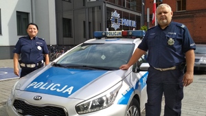 Dwoje umundurowanych policjantów stoi przy radiowozie na tle komendy
