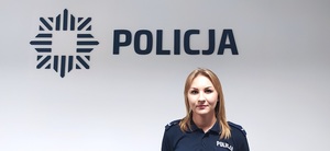 Umundurowana policjantka stoi na tle ściany z logiem i napisem POLICJA
