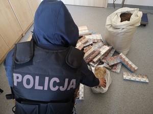 Nieumundurowany policjant kuca przed leżącymi na podłodze paczkami papierosów i suszem tytoniowym