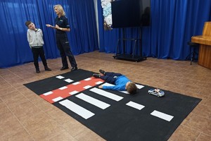 Chłopczyk leżący na macie imitującej przejście dla pieszych oraz stojąca policjantka z innym chłopczykiem.
