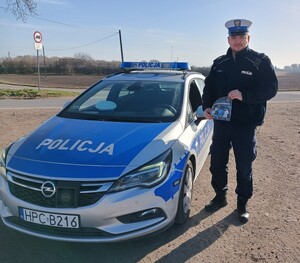 Policjant stoi przy radiowozie z ulotką "Łapki na kierownicę"