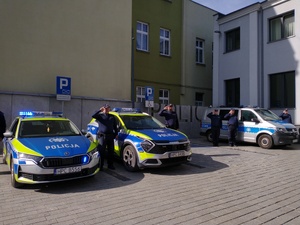 Umundurowani policjanci na tle radiowozów z włączonymi sygnałami oddają hołd poległemu koledze.